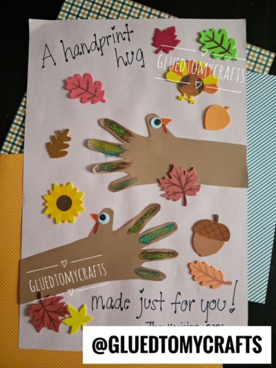A handmade craft on light pink paper featuring two turkey handprints with glittered fingers, surrounded by foam leaf, sunflower, and acorn stickers, with the text A handprint hug made just for you!.
