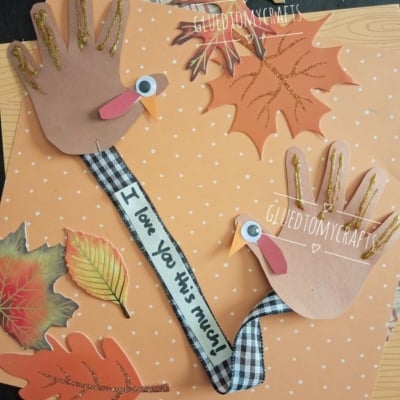Two handprint turkeys made of brown paper, googly eyes, and ribbon are surrounded by decorative autumn leaves on an orange polka dot background. One ribbon says I love you this much..