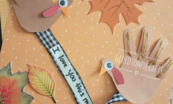 Two handprint turkeys made of brown paper, googly eyes, and ribbon are surrounded by decorative autumn leaves on an orange polka dot background. One ribbon says I love you this much..