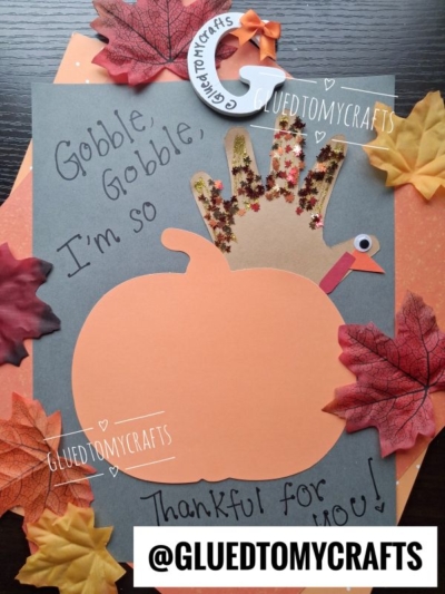 A Thanksgiving craft features a handprint turkey and a large paper pumpkin, surrounded by faux autumn leaves. Perfect for anyone searching for FALL CRAFTS FOR KIDS, the display says, “Gobble, gobble, I’m so thankful for you!” and is labeled @GluedToMyCrafts.
