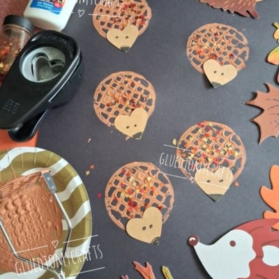 Brown paper hedgehog crafts with fall leaf confetti are arranged on a dark paper background. Crafting supplies like glue, a paper punch, and paint sit nearby. The @GLUEDTOMYCRAFTS watermark is visible.