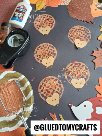 Brown paper hedgehog crafts with fall leaf confetti are arranged on a dark paper background. Crafting supplies like glue, a paper punch, and paint sit nearby. The @GLUEDTOMYCRAFTS watermark is visible.