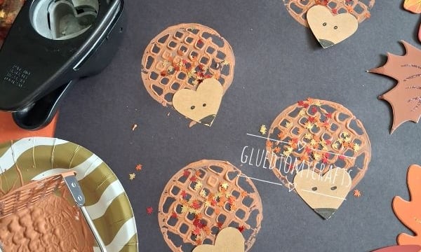 Brown paper hedgehog crafts with fall leaf confetti are arranged on a dark paper background. Crafting supplies like glue, a paper punch, and paint sit nearby. The @GLUEDTOMYCRAFTS watermark is visible.