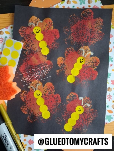 Caterpillars made from yellow dot stickers with smiley faces are on a black paper with orange leaf prints, making this scene perfect for FALL CRAFTS FOR KIDS. Art supplies and fall-themed paper surround the artwork.