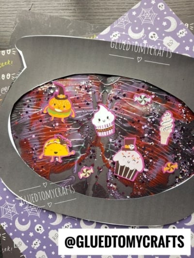 Cat-shaped Halloween craft decorated with cute stickers—cupcakes, a donut, and candies—on a purple and black background with stars, webs, bats, and paint splat crafts. @GLUEDTOMYCRAFTS is printed at the bottom.