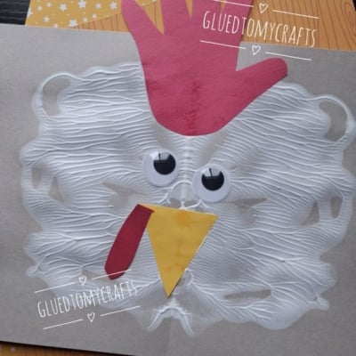 A chicken craft made from a handprint red comb, paper beak and wattle, googly eyes, and white paint spread in a circle, displayed on patterned paper. @GLUEDTOMYCRAFTS is written on the photo.