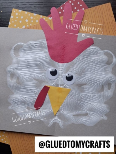 A chicken craft made from a handprint red comb, paper beak and wattle, googly eyes, and white paint spread in a circle, displayed on patterned paper. @GLUEDTOMYCRAFTS is written on the photo.