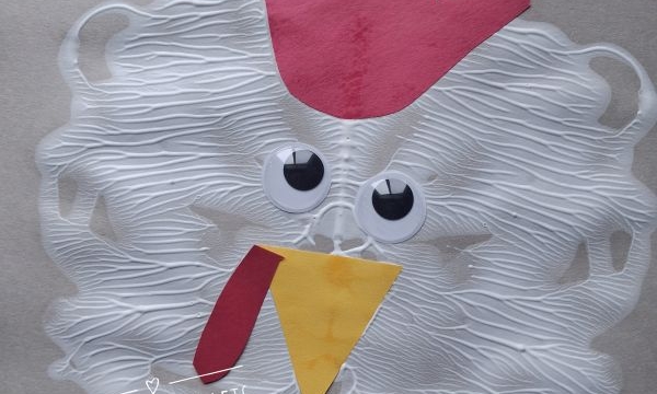 A chicken craft made from a handprint red comb, paper beak and wattle, googly eyes, and white paint spread in a circle, displayed on patterned paper. @GLUEDTOMYCRAFTS is written on the photo.