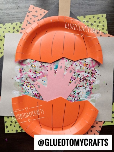 A pumpkin craft made from an orange paper plate, with a pink paper hand inside and colorful glitter, features a paint splat exploding pumpkin design on a white background. Craft supplies and patterned paper are visible underneath.