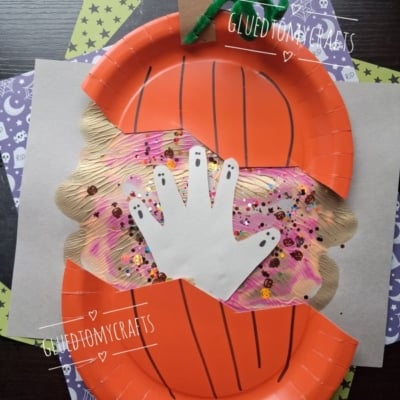 A paper plate pumpkin craft with a handprint cutout in the center, decorated with sequins and glitter on colorful paper, sits on a stack of patterned papers. @GLUEDTOMYCRAFTS is written at the bottom.