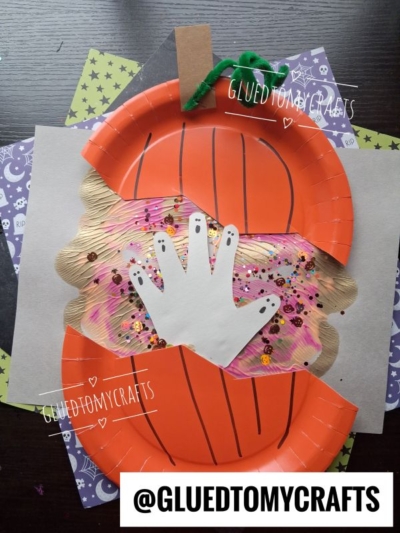 A paper plate pumpkin craft with a handprint cutout in the center, decorated with sequins and glitter on colorful paper, sits on a stack of patterned papers. @GLUEDTOMYCRAFTS is written at the bottom.