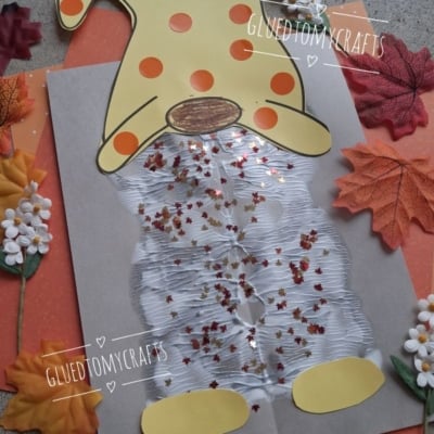 A fall gnome craft made with paper, cotton, glitter, and sequins. The gnome has an orange hat with yellow dots and is surrounded by artificial leaves and flowers. @GLUEDTOMYCRAFTS is labeled on the image.