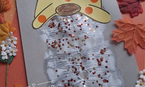 A fall gnome craft made with paper, cotton, glitter, and sequins. The gnome has an orange hat with yellow dots and is surrounded by artificial leaves and flowers. @GLUEDTOMYCRAFTS is labeled on the image.