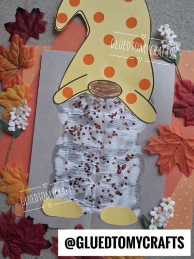 A paper craft gnome with a yellow hat and orange spots, white beard, and oval nose, surrounded by fake autumn leaves and white flower decorations. The hat reaches the top left corner.