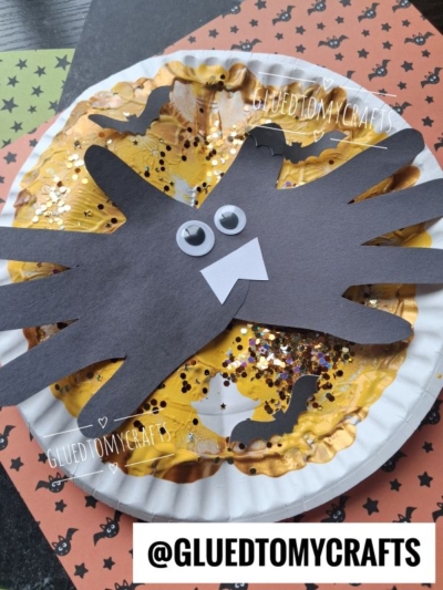 A paper plate with gold paint, glitter, and black bat cutouts; a black paper spider with googly eyes and fangs is on top. Halloween-themed background. Text: @GLUEDTOMYCRAFTS.