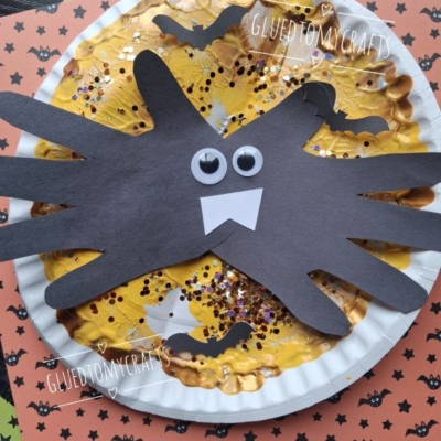 Paper plate spider craft with black handprint legs, googly eyes, and white fangs on a yellow, glittery plate. Background features orange and black bat-themed paper and the text @GLUEDTOMYCRAFTS.