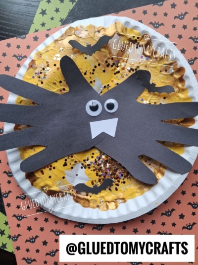 Paper plate spider craft with black handprint legs, googly eyes, and white fangs on a yellow, glittery plate. Background features orange and black bat-themed paper and the text @GLUEDTOMYCRAFTS.