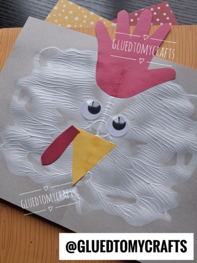 A paper chicken craft with a handprint red comb, googly eyes, a yellow beak, and swirled white paint feathers on gray paper. Scrapbook paper is in the background. Has @GLUEDTOMYCRAFTS watermark.
