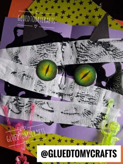 A cat face made from white tape, green cat eyes, black paper ears, and set on purple and yellow star paper resembles a playful paint splat mummy. Small colorful plastic skeletons are on the sides. @GLUEDTOMYCRAFTS watermark is present.