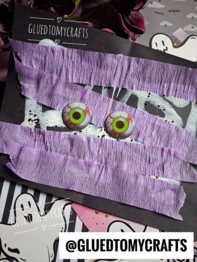 A spooky paint splat craft featuring purple crepe paper strips, two green eyeball decorations, and ghost-themed paper in the background. The username @GLUEDTOMYCRAFTS appears on the image.