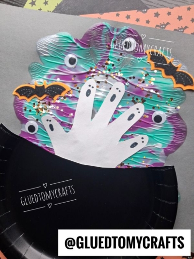A Halloween craft featuring a paper handprint ghost surrounded by googly eyes, glitter, and small bat shapes, partly tucked into a cut black paper plate. The username @gluedtomycrafts is displayed.