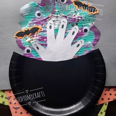 A paper craft shows a black cauldron with handprint ghosts, bats, and colorful swirls rising from it, on a gray background. The handle @GLUEDTOMYCRAFTS is written at the bottom.
