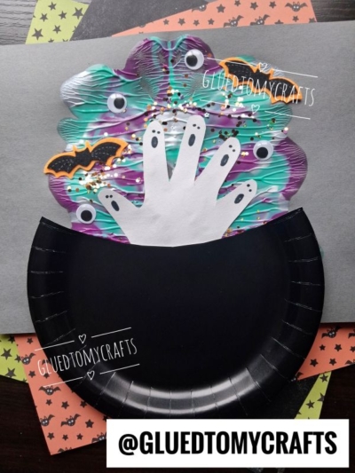 A paper craft shows a black cauldron with handprint ghosts, bats, and colorful swirls rising from it, on a gray background. The handle @GLUEDTOMYCRAFTS is written at the bottom.