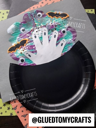A black paper plate cauldron with colorful foam above it, decorated with cutout handprints as ghosts, bats, sequins, and star-shaped confetti. The username @GLUEDTOMYCRAFTS is displayed.