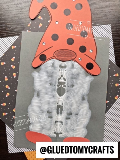 A paint splat gnome with an orange polka-dot hat and feet is placed over a skeleton X-ray image on Halloween-themed paper. @GLUEDTOMYCRAFTS is written on the image.