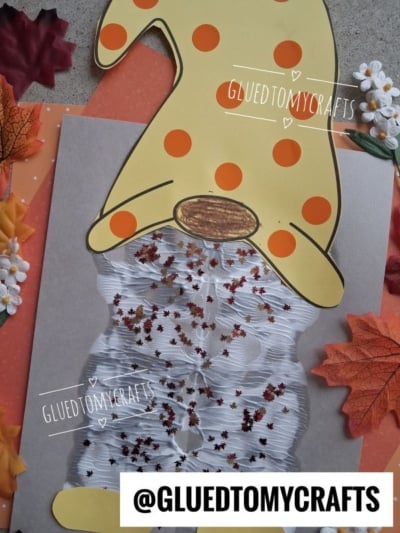 A fall gnome craft made with a yellow and orange polka-dot hat, white crinkled beard with leaf-shaped confetti, surrounded by fake autumn leaves and flowers. The username @GLUEDTOMYCRAFTS is visible.