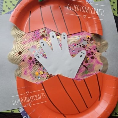 A paper plate pumpkin craft with a cut-out handprint ghost in the center, decorated with sequins and glitter, on a Halloween-themed background. @GLUEDTOMYCRAFTS text appears on the image.