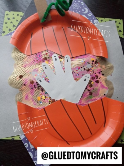 A paper plate pumpkin craft with a cut-out handprint ghost in the center, decorated with sequins and glitter, on a Halloween-themed background. @GLUEDTOMYCRAFTS text appears on the image.