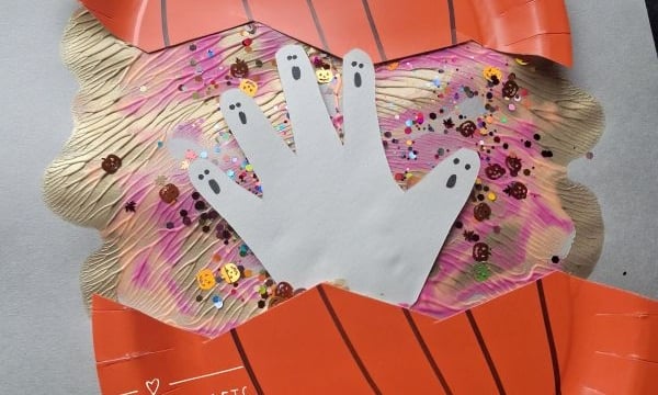 A paper plate pumpkin craft with a cut-out handprint ghost in the center, decorated with sequins and glitter, on a Halloween-themed background. @GLUEDTOMYCRAFTS text appears on the image.