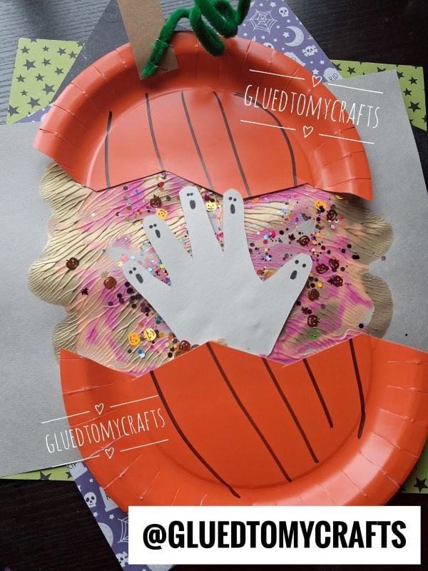 A paper plate pumpkin craft with a cut-out handprint ghost in the center, decorated with sequins and glitter, on a Halloween-themed background. @GLUEDTOMYCRAFTS text appears on the image.
