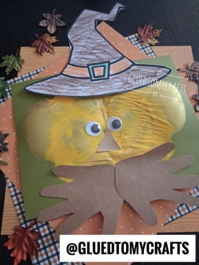 A handmade scarecrow face craft with a witch hat, large googly eyes, a yellow painted face, and brown paper hands. Autumn leaves and plaid paper decorate the background. @GLUEDTOMYCRAFTS is written at the bottom.