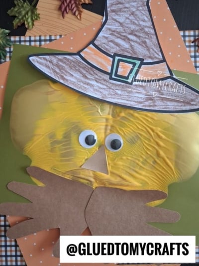 A Halloween craft features a yellow scarecrow face with googly eyes, brown paper hands, and a large witch hat on a fall-themed background. The image has the text @GLUEDTOMYCRAFTS at the bottom.