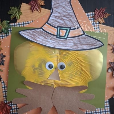 A fall-themed craft of a scarecrow face made from a yellow paint-filled bag, googly eyes, paper hat, and hands, on a green background with autumn leaves and orange plaid paper. @GLUEDTOMYCRAFTS text at the bottom.