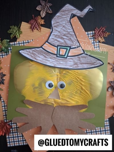 A fall-themed craft of a scarecrow face made from a yellow paint-filled bag, googly eyes, paper hat, and hands, on a green background with autumn leaves and orange plaid paper. @GLUEDTOMYCRAFTS text at the bottom.
