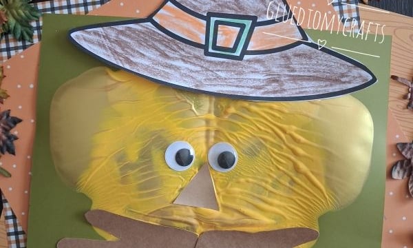A fall-themed craft of a scarecrow face made from a yellow paint-filled bag, googly eyes, paper hat, and hands, on a green background with autumn leaves and orange plaid paper. @GLUEDTOMYCRAFTS text at the bottom.
