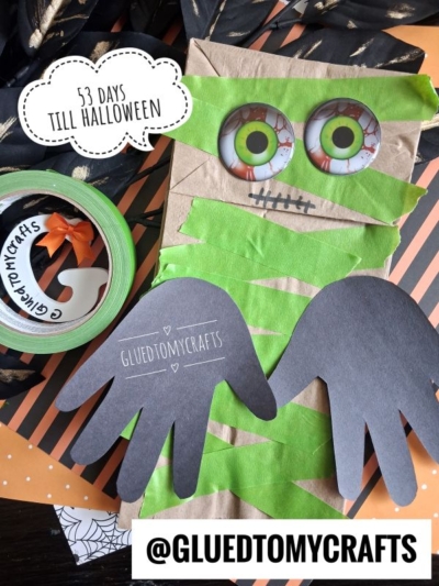 A fun Halloween Crafts for Kids project: a paper bag mummy craft with googly eyes, green tape, and black paper hands. A sticker says “53 days till Halloween” and a tag reads @GLUEDTOMYCRAFTS. Craft tape and scissors are also visible.