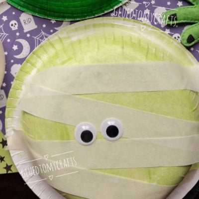A green paper bowl mummy made from a plate decorated with masking tape strips and googly eyes.
