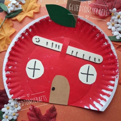 A paper plate painted red resembles an apple, decorated with paper cutouts for windows, a door, a popsicle stick roof, and a green leaf, surrounded by fake autumn leaves. @GluedToMyCrafts appears on the image.