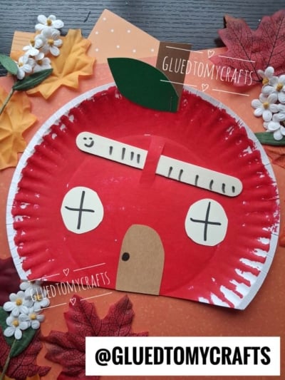 A paper plate painted red resembles an apple, decorated with paper cutouts for windows, a door, a popsicle stick roof, and a green leaf, surrounded by fake autumn leaves. @GluedToMyCrafts appears on the image.