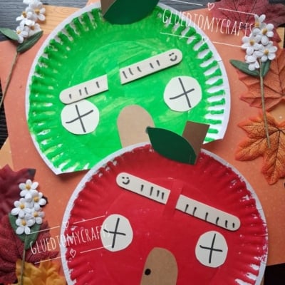 Two paper plate apples, one green and one red, are decorated with paper stems, leaves, and silly faces. The plates are surrounded by artificial flowers and autumn leaves on a wooden surface.