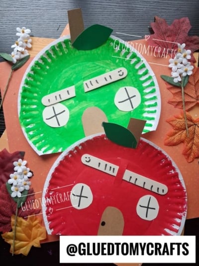 Two paper plate apples, one green and one red, are decorated with paper stems, leaves, and silly faces. The plates are surrounded by artificial flowers and autumn leaves on a wooden surface.