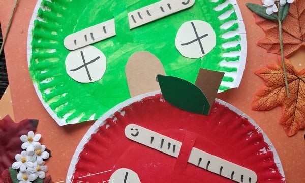 Two paper plate apples, one green and one red, are decorated with paper stems, leaves, and silly faces. The plates are surrounded by artificial flowers and autumn leaves on a wooden surface.