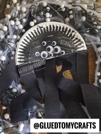 A black paper plate spider craft with googly eyes and black streamers for legs, surrounded by faux cobwebs and white pom-poms—perfect for Halloween Crafts for Kids. @GLUEDTOMYCRAFTS text appears at the bottom.