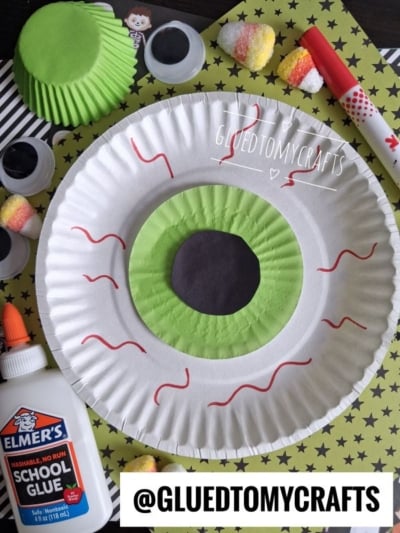 A large paper plate decorated as a green eyeball with red veins, surrounded by craft supplies like glue, googly eyes, green cupcake liners, a marker, and candy—perfect inspiration for Halloween Crafts for Kids. @GLUEDTOMYCRAFTS is written on the image.