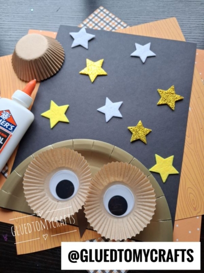 A paper owl made from cupcake liners and googly eyes sits on a black paper with yellow and white star stickers—an adorable inspiration for FALL CRAFTS FOR KIDS. Glue and craft paper are nearby, ready for more creative fun. The username @GLUEDTOMYCRAFTS is at the bottom.