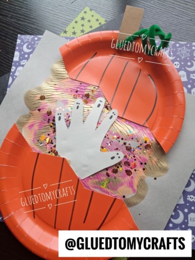 A pumpkin craft made from orange paper plates, glitter, and a white handprint cutout, placed on Halloween-themed paper. Green pipe cleaner acts as the stem. Text: @GLUEDTOMYCRAFTS.